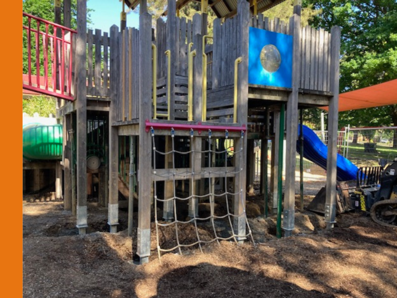 The image shows a wooden playground structure with multiple levels, a rope climbing wall, and slides. The structure is surrounded by a mulch-covered ground, and a fence can be seen in the background, suggesting the area may be under maintenance or renovation. An orange shade cloth provides partial covering overhead, and some playground equipment is visible, including a green tunnel slide and a blue slide.