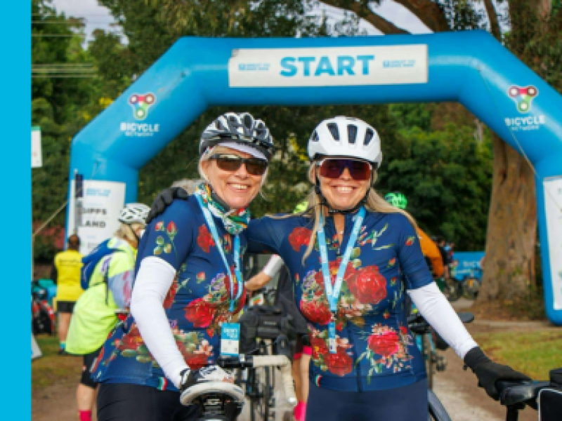 Two smiling cyclists wearing matching floral jerseys stand together near the starting line of a cycling event. They are wearing helmets, sunglasses, and medals around their necks. In the background, a blue inflatable arch marked 'START' is visible, along with other participants preparing for the event.