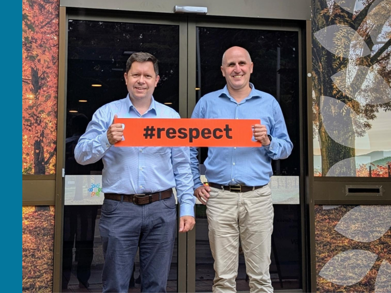  Two men standing in front of a building with autumn-themed window panels, both wearing light blue shirts and holding an orange sign with the hashtag '#respect'.