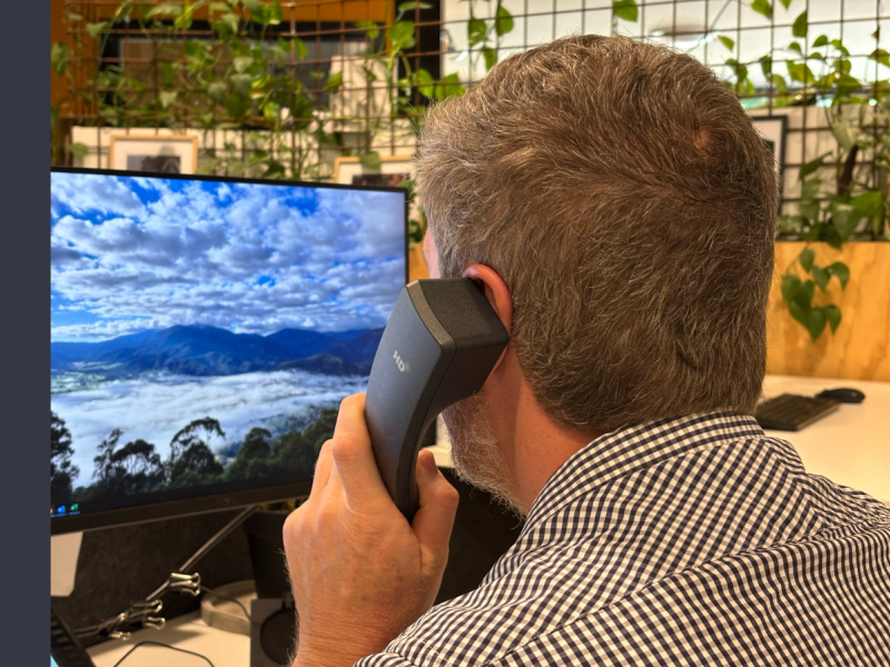 Man in a checkered shirt holding a black landline phone to his left ear whilst looking at a screen with an image of mountains and clouds as the screen saver.