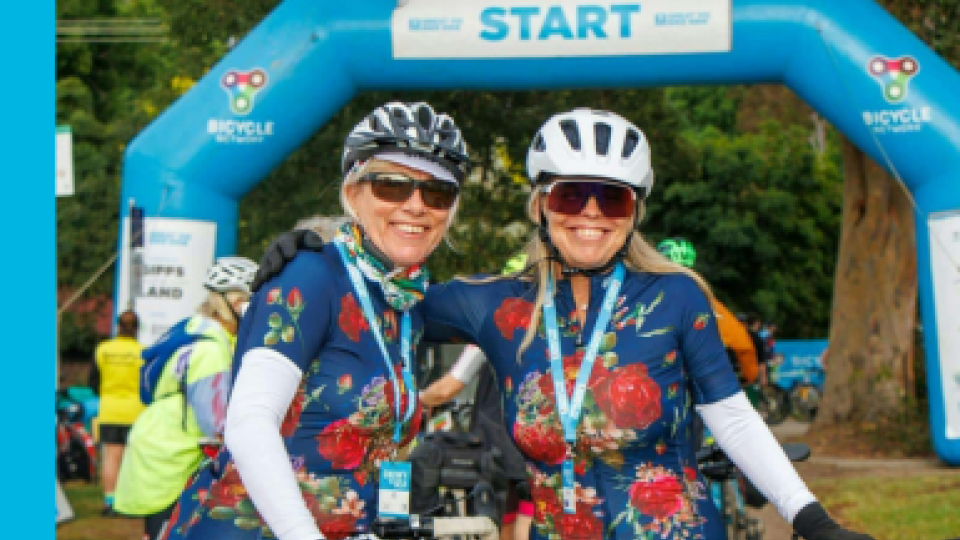 Two smiling cyclists wearing matching floral jerseys stand together near the starting line of a cycling event. They are wearing helmets, sunglasses, and medals around their necks. In the background, a blue inflatable arch marked 'START' is visible, along with other participants preparing for the event.